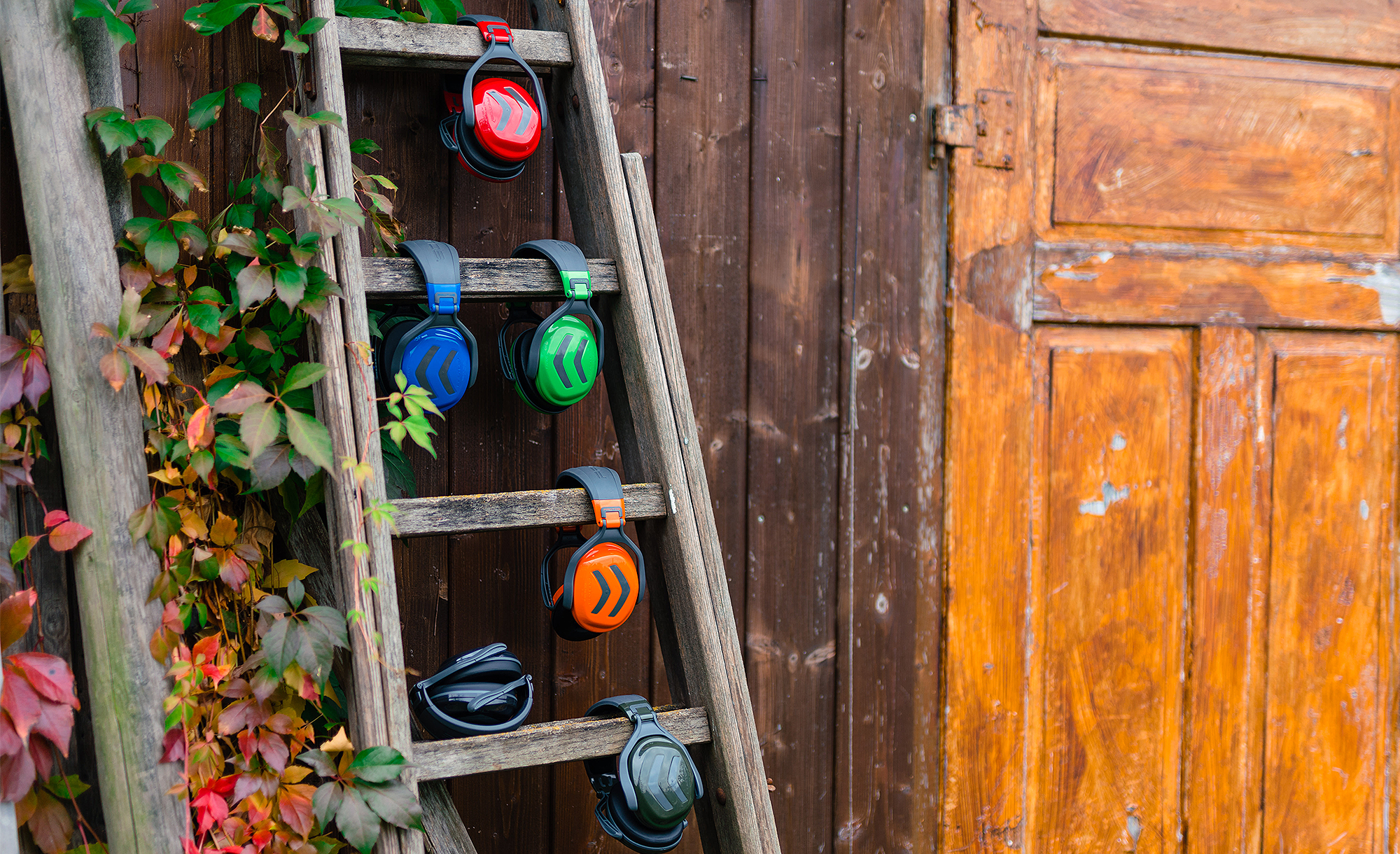 Zes kleurrijke gehoorbeschermers hangen aan een oude houten ladder tegen een houten wand met herfstbladeren.