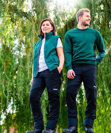 Twee personen in groene tuinkleding staan naast elkaar voor een achtergrond met hangende takken.