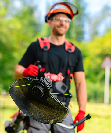 Man met helm en veiligheidsbril houdt een bosmaaier met draadkop vast, met groen landschap op de achtergrond.