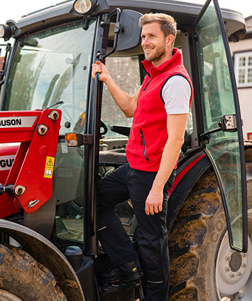 Man met rood vest stapt in een tractor met open cabinedeur.