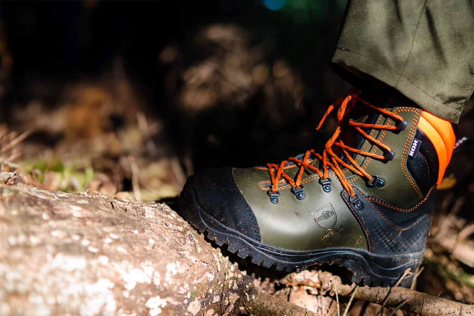 Close-up van een robuuste boslaars in groen en zwart met oranje veters. De schoen staat op een boomstam in het bos, met op de achtergrond een onscherpe bosbodem en takken.