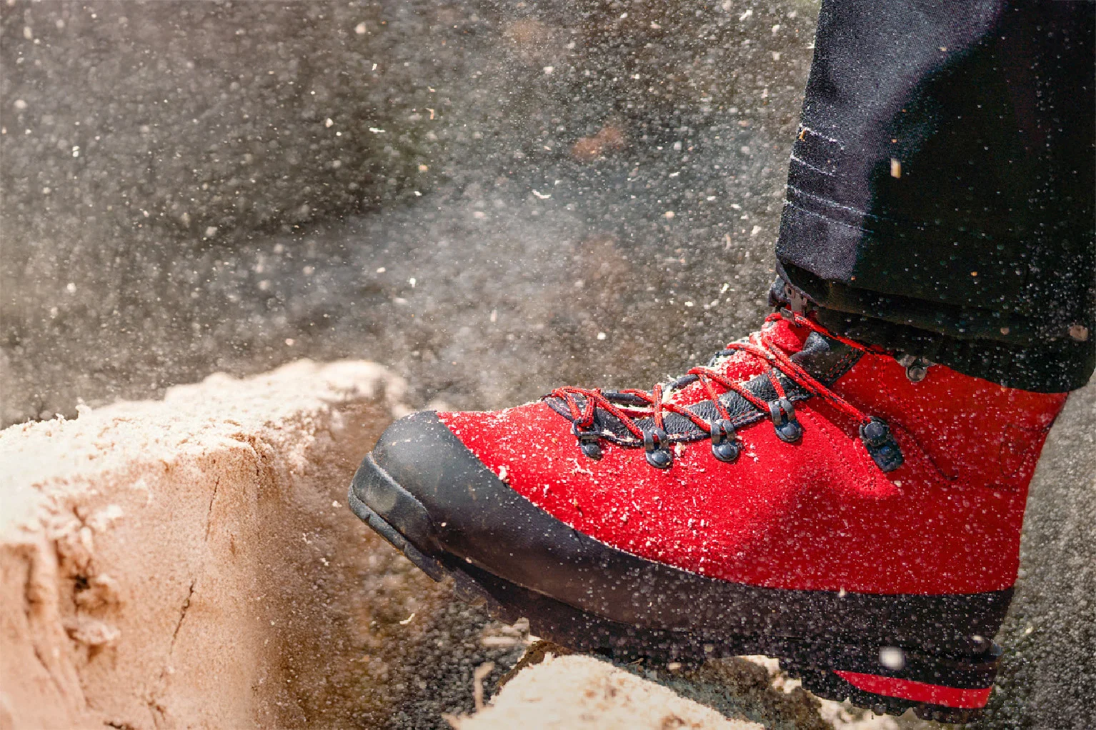 Close-up van een robuuste boslaars in groen en zwart met oranje veters. De schoen staat op een boomstam in het bos, met op de achtergrond een onscherpe bosbodem en takken.