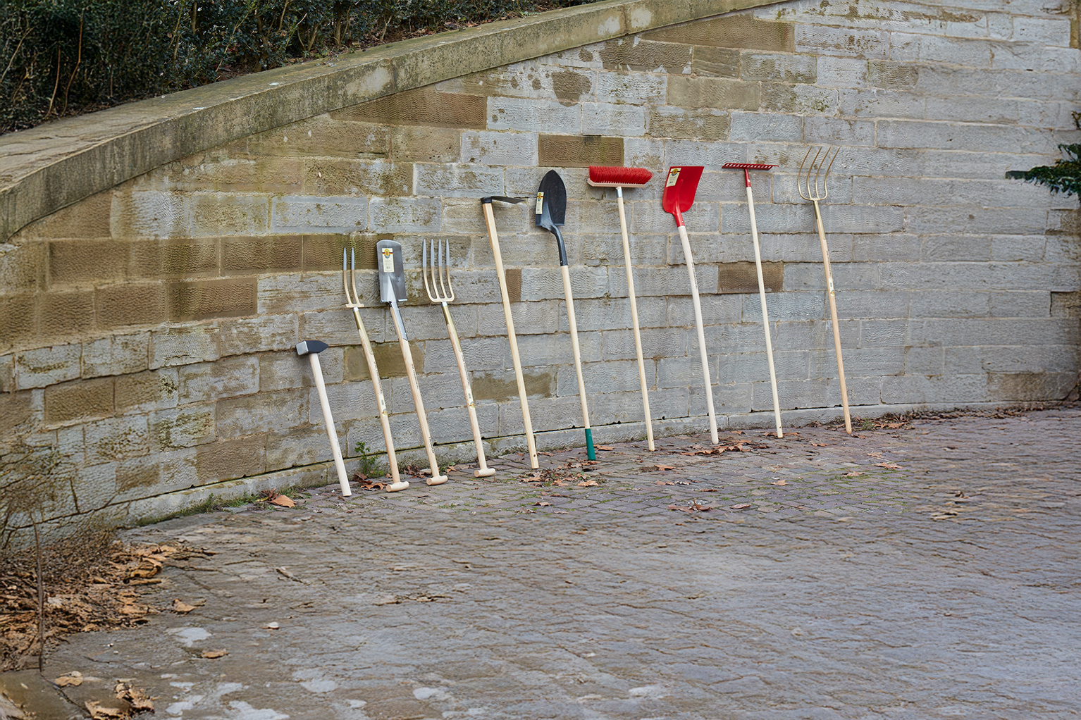 Meerdere tuingereedschappen met houten stelen staan tegen een stenen muur, waaronder een hamer, spitvorken, een schop, een bezem, een sneeuwschop, een hark en een riek.