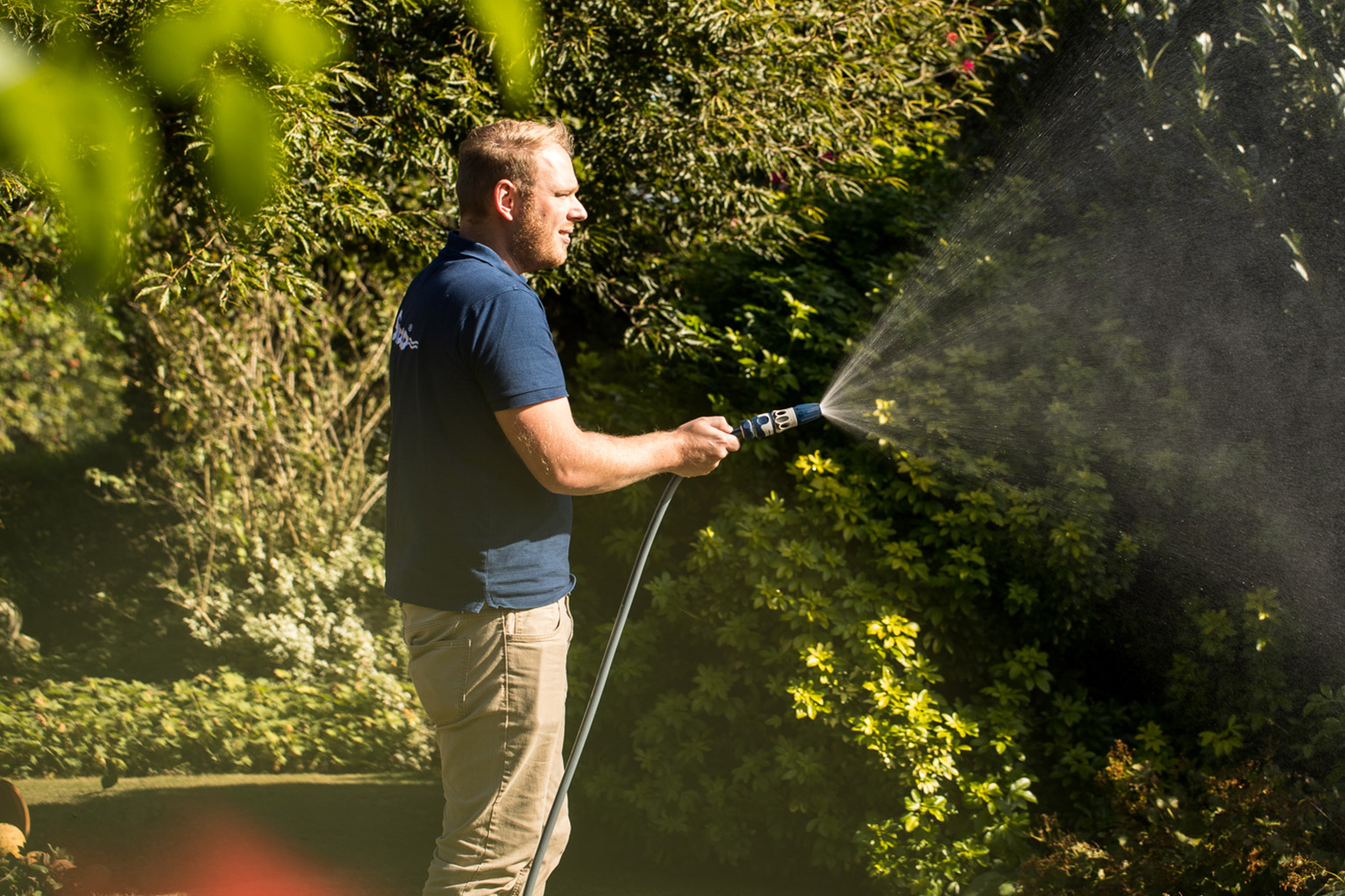Een man staat in de tuin en besproeit met een sproeikop dichte struiken, waarbij de fijne waternevel goed zichtbaar is in het zonlicht.