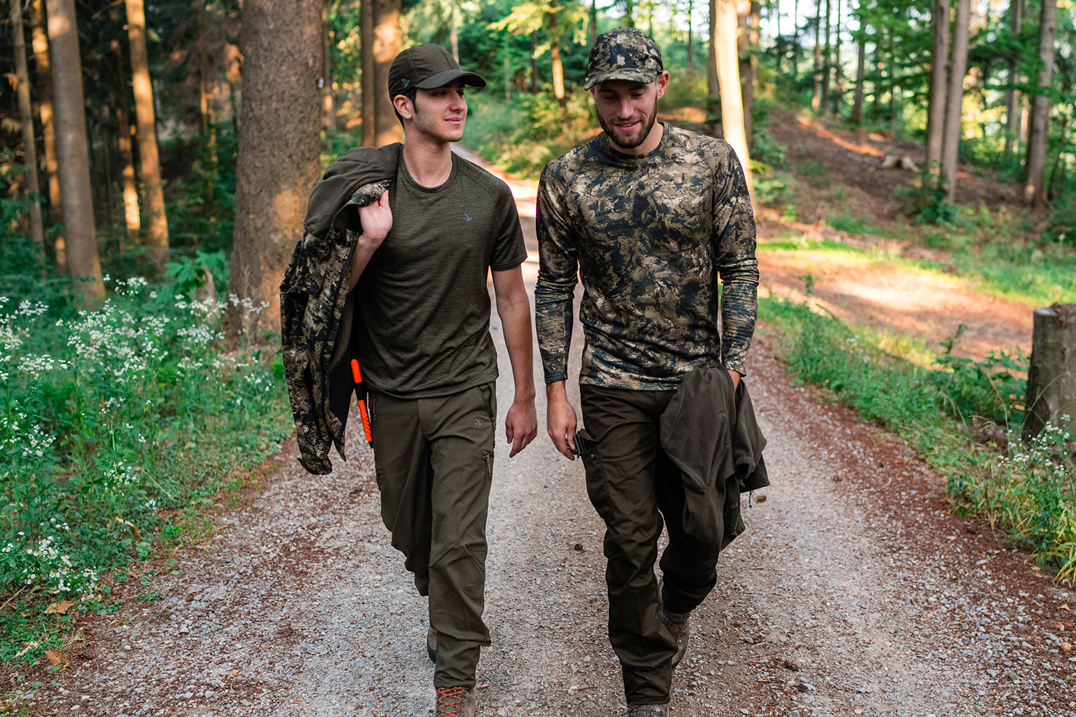 Twee mannen in functionele outdoor- of jachtkleding lopen naast elkaar op een bospad; dragen petten, jassen in de hand en zijn in gesprek.