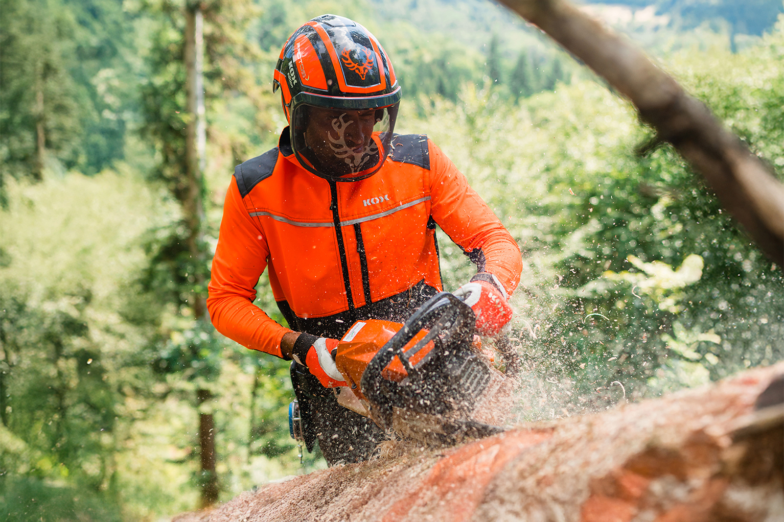 Een persoon in oranje bosbouwbeschermkleding en met helm zaagt in het bos met een kettingzaag aan een boomstam, terwijl zaagsel door de lucht vliegt.