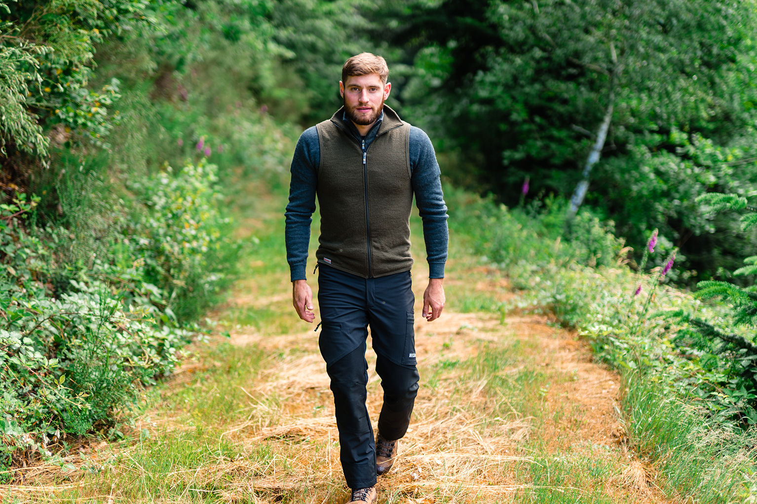 Een man loopt op een bospad, omgeven door groen bos. Hij draagt een fleecetrui, outdoorbroek en wandelschoenen.