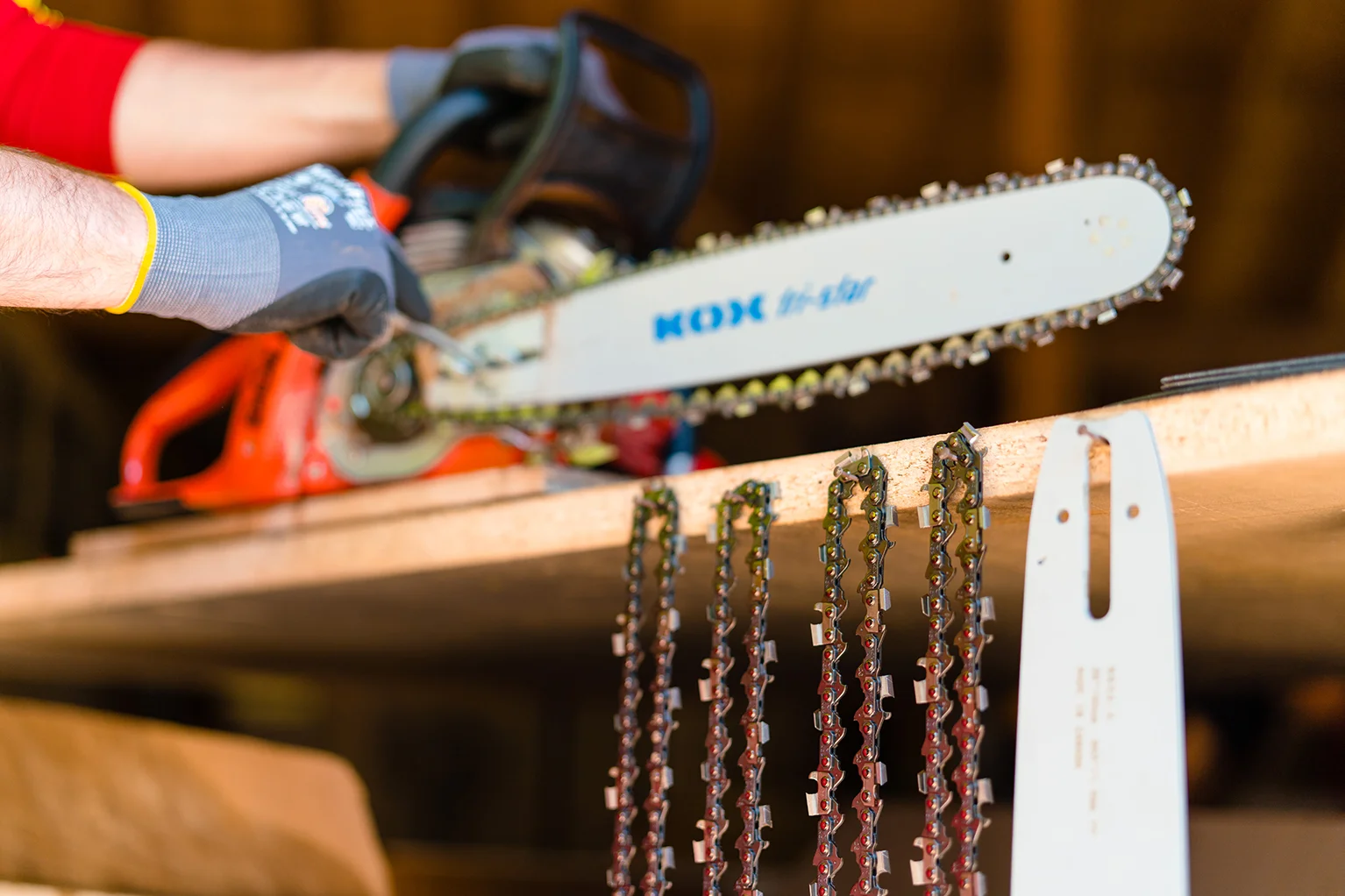 Persoon met handschoenen spant een KOX zaagketting op een kettingzaag, op de voorgrond hangen meerdere reservekettingen en een zaagblad aan een plank.