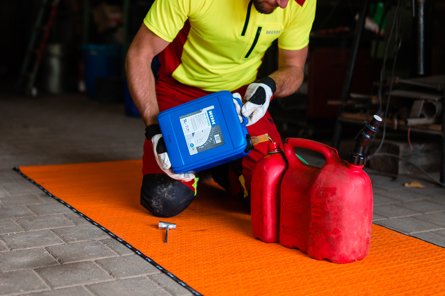 Een persoon in opvallende beschermende kleding giet in een werkplaats vloeistof uit een blauwe jerrycan in twee rode brandstofcontainers.